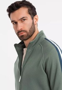 Chaqueta de forro polar verde con cremallera y cuello alto, que presenta franjas laterales en azul marino y blanco, además de una textura suave. La cremallera es metálica.