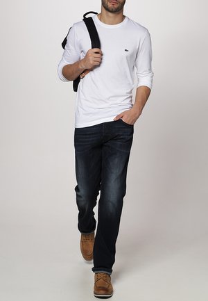 T-shirt blanc à manches longues avec un petit logo vert, associé à un jean bleu foncé et des chaussures marron. Un homme portant un sac à dos noir.