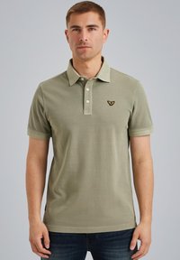 Polo en vert olive, fabriqué en coton, doté d'une patte de boutonnage à trois boutons, de manches courtes et d'un logo brodé sur la poitrine.