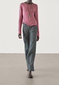 Pull rose à manches longues, coupe ample, associé avec un pantalon large rayé gris clair. Le modèle porte des sandales noires et un collier circulaire.