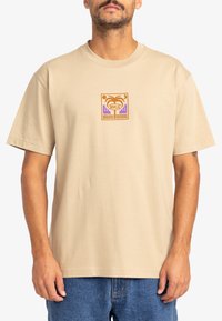 Beige cotton t-shirt featuring a square graphic with a palm tree, buildings, and waves in orange and purple tones at the center. Short sleeves.