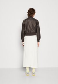 Hnědá kožená bomber bunda s žebrovanými manžetami, doplněná dlouhou bílou sukní. Nosič má na sobě žluté ponožky a tenisky s šedými a žlutými akcenty.