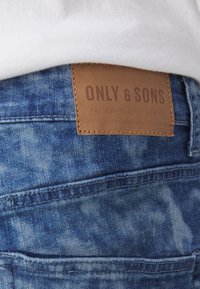 Modré džínové kalhoty s texturovaným povrchem; obsahují hnědou koženou nášivku s nápisem "ONLY & SONS" a "PASSIONATE JEANS MAKERS."