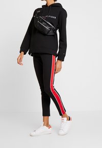 Sudadera negra con logo, pantalones negros a juego con rayas laterales rojas y blancas, y zapatillas blancas, complementados con un bolso cruzado negro.