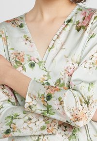 Top avvolgente floreale con scollatura a V; realizzato in tessuto morbido e leggero con motivi floreali verdi, rosa e bianchi; maniche lunghe e fluide.