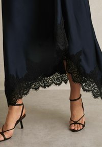 Abito di raso blu navy con un intricato bordo in pizzo nero lungo l'orlo, caratterizzato da bordi frangiati. Tacchi neri con cinturini completano il look.