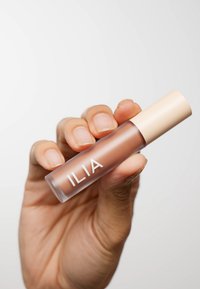 ILIA Lipgloss in einer klaren Tube mit einem beigen Deckel, der eine abgerundete Form und glatte Textur aufweist. Das Produktetikett zeigt "ILIA" in Weiß.
