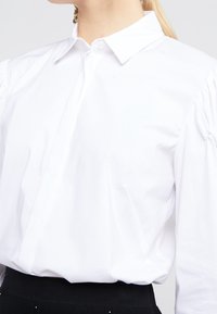 Camicia bianca a maniche lunghe con colletto classico, tessuto texturizzato e dettagli sulle spalle a sbuffo, abbinata a un capo scuro in vita.