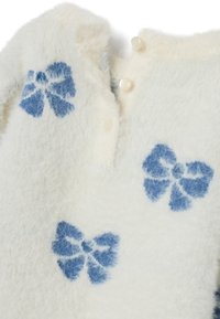 Maglione bianco peloso con motivi a fiocco blu. Presenta una scollatura con tre bottoni bianchi. Tessuto morbido.