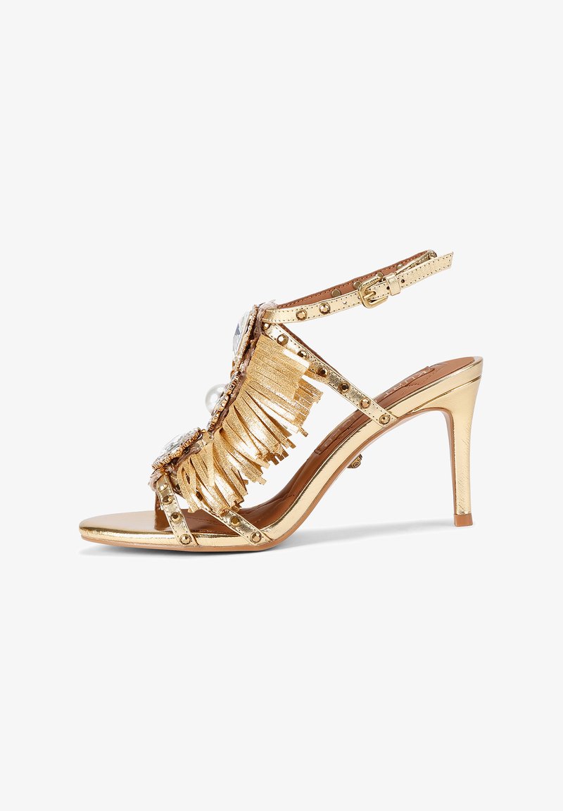 Złota sandałka na wysokim obcasie z frędzlami, dużymi ozdobnymi kamieniami i paskiem z klamrą. Błyszczący materiał z detalami w postaci ćwieków. Otwarty przód.