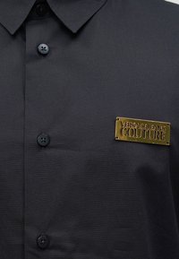 Μαύρο πουκάμισο με κουμπιά και χρυσή μεταλλική πλάκα στο στήθος με την επιγραφή "Versace Jeans Couture."