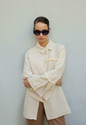 Γυναίκα που φοράει oversized μπεζ παλτό με κουμπιά, φαρδιά μανίκια και μεγάλα σκούρα γυαλιά ηλίου στέκεται μπροστά σε έναν απλό φωτεινό τοίχο με τα χέρια σταυρωμένα.