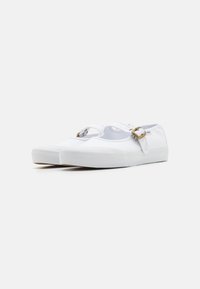 Zapatillas de lona blancas con suela de goma, que cuentan con dos correas ajustables y hebillas de metal de color dorado. Textura suave y forma redondeada.