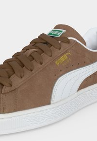 Καφέ σουέτ αθλητικό Puma με λευκή σόλα και πλαϊνή λωρίδα, χρυσό λογότυπο Puma Suede, καφέ κορδόνια και πράσινη ετικέτα στη γλώσσα.