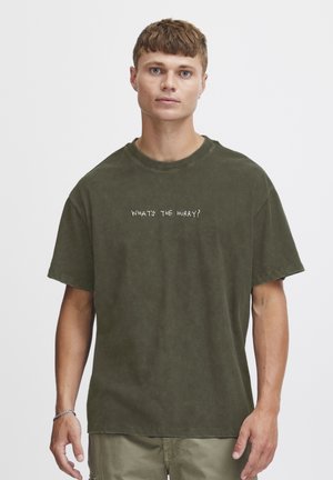 Olive green oversized t-shirt in soft cotton with a round neck, featuring white text "WHAT'S THE HURRY?" on the front. Short, wide sleeves.