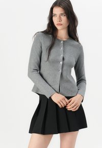 Grå strikket cardigan med rund hals, frontknap lukning og sølvknapper, parret med en sort plisseret nederdel.
