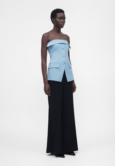 Γυναίκα που φοράει αμάνικη ανοιχτό μπλε μπλούζα με κουμπιά και τσέπες με καπάκια και μαύρο παντελόνι με φαρδιά πόδια, στέκεται μπροστά σε λευκό φόντο.