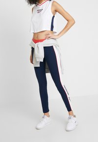 Kvinna som modellerar en vit Reebok crop top, marinblå leggings med vita sidostrimmor, vita sneakers och en grå sweatshirt knuten runt midjan.