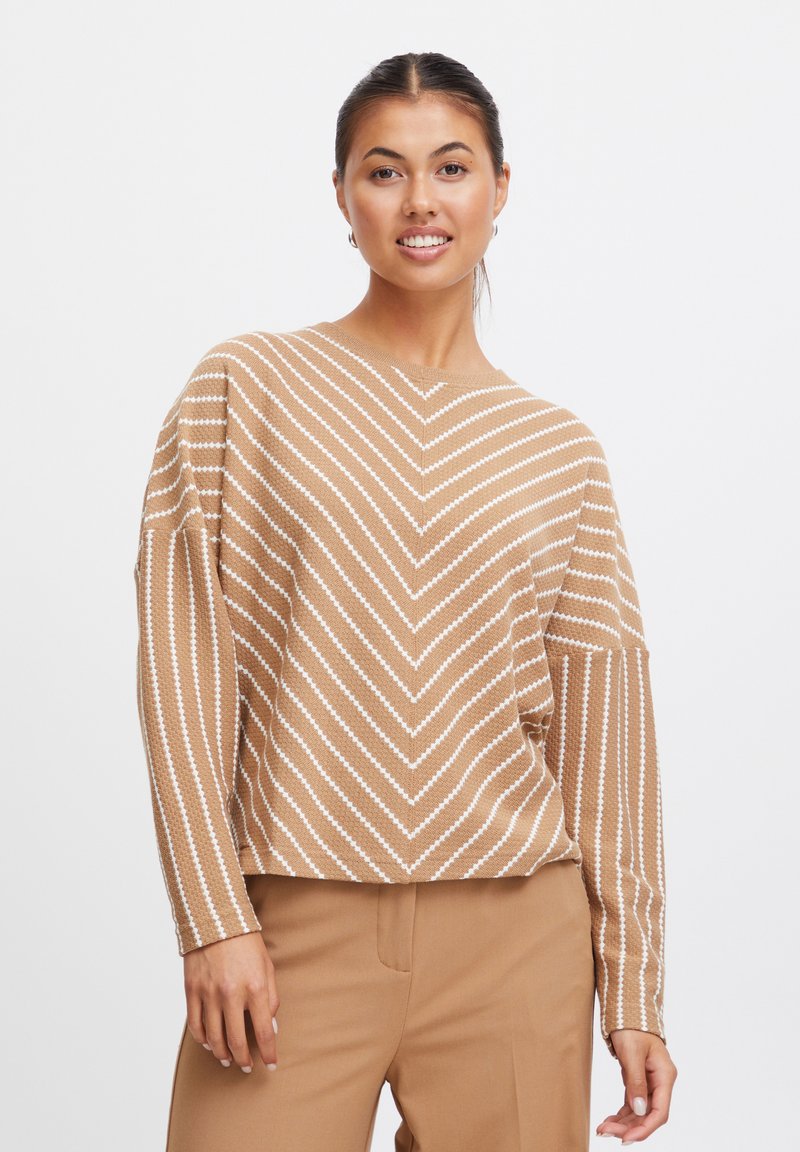 Pull en maille marron clair avec des rayures en chevron blanches, col rond et épaules tombantes. Associé à un pantalon beige.