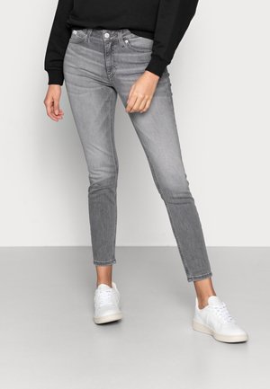 Person wearing slim-fit, faded gray jeans paired with white sneakers and a black long-sleeve top, standing against a plain background.