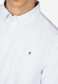 Camisa de botones azul claro con rayas blancas, que presenta un pequeño logo negro en el lado izquierdo del pecho y un diseño de cuello clásico.