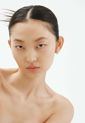 Primer plano de una persona con cabello negro lacio. Tiene una piel suave, labios neutros y un pequeño pendiente brillante en la oreja izquierda.