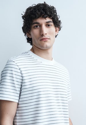 Ung mand med krøllet mørkt hår iført en hvid T-shirt med tynde sorte horisontale striber mod en ensartet lys baggrund.