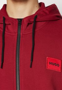Červená zipová mikina s texturovanou tkaninou. Obsahuje černý zip a šňůrky, s černým logem "HUGO" na obdélníkovém červeném nápletu.