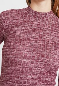 Burgundowy sweter dzianinowy z teksturowanym wzorem, o wysokiej stójce i subtelnym rozciągnięciu. Dopasowany krój podkreśla całościowy design.