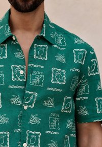 Green linen short-sleeve shirt with white leaf and frame patterns, button-up front, and a classic collar. Textured fabric detail visible.