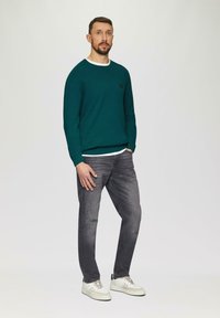 Türkisfarbener Strickpullover mit weißem Kragen, graue distressed Jeans und cremefarbene Sneaker mit Schnürsenkeln und seitlichen Streifen. Model steht vor einem neutralen Hintergrund.