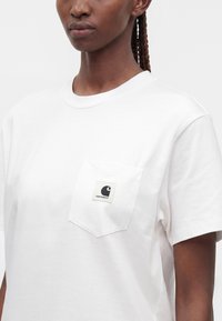 Γυναίκα που φοράει απλό λευκό T-shirt με κοντά μανίκια και τσέπη στο στήθος με μικρό λογότυπο της Carhartt.