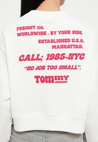 Witte sweatshirt met roze grafische tekst op de achterkant, met de merknaam "TOMMY Jeans" en verschillende zinnen in vette letters.