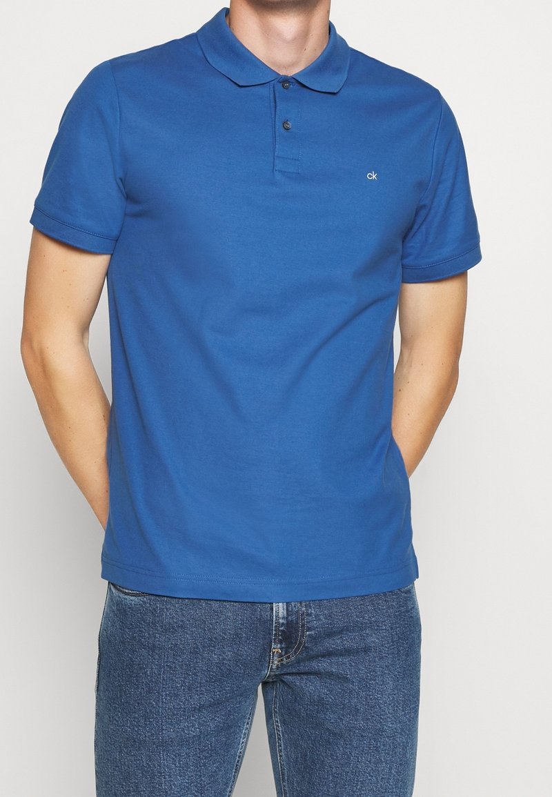 Homme portant un polo bleu à manches courtes avec un petit logo blanc "ck" et un jean bleu, les mains dans les poches.