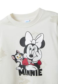 Minnie Mouse avec un grand nœud et une robe rouge, souriante avec un chat noir et blanc à ses côtés, au-dessus du texte "MINNIE" sur un tissu blanc.