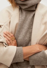 Manteau beige sur un col roulé côtelé gris, avec les mains croisées et portant une bague en or. Textures douces et palette de couleurs neutres.