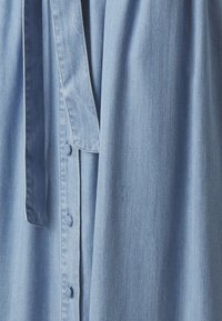 Vestido de mezclilla azul claro con botones en el frente, cintura anudada y tejido suave, ligeramente texturizado. Diseño casual distintivo.