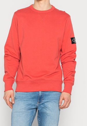 Uomo che indossa un felpa a girocollo rosso corallo con patch del logo Calvin Klein sulla manica sinistra e jeans azzurri, in piedi contro uno sfondo neutro.