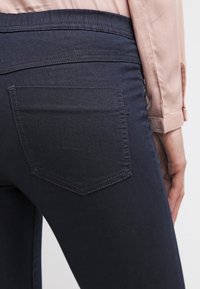 Tummansiniset farkut, joissa on istuva siluetti, varustettuna tavallisella läppätaskulla ja sileällä pinnalla. Vyötärö on hieman poimutettu.