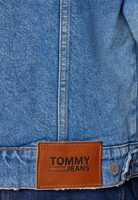 Blå denimjacka med kontrastsömmar, en brun läderetikett präglad med "TOMMY JEANS" och metalliska knappdetaljer.