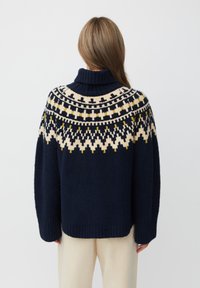 Granatowy sweter robiony na szydełku z wysokim kołnierzem, z wzorzystymi akcentami w kolorze kremowym i żółtym na karczku oraz luźnym, teksturowanym materiałem.