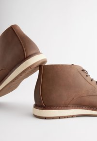 Stivaletti alla caviglia in pelle marrone con una texture liscia, dotati di suola in gomma color crema, design con lacci e dettagli di cuciture a contrasto.