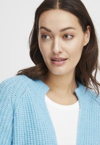 Cardigan lavorato a maglia azzurro chiaro con motivo testurizzato e collo a coste, indossato sopra una camicia bianca, caratterizzato da un design con apertura frontale.