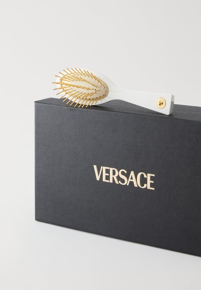 Λευκή βούρτσα μαλλιών με χρυσές τρίχες, με λαβή με υφή και στρογγυλό σχήμα, τοποθετημένη πάνω σε μια υφασμένη μαύρη θήκη με την επιγραφή "VERSACE" σε χρυσό.