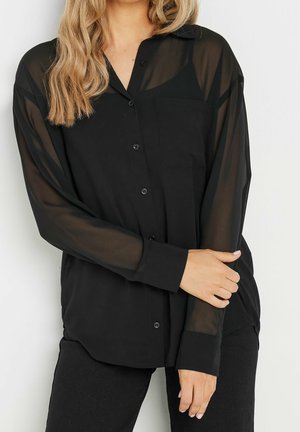 Nainen, joka on pukeutunut mustaan läpikuultavaan napitettuun paitapuseroon, jossa on etutasku ja pitkät hihat, yhdistettynä mustiin housuihin, seisoo yhdellä käsivarrella ristissä.