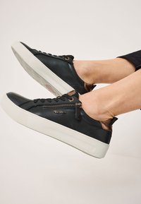 Sneakers en cuir noir avec une semelle en caoutchouc blanche, bout rond, accents bruns contrastants et zips décoratifs sur les côtés.