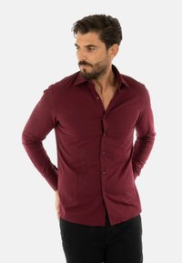 Camicia a maniche lunghe bordeaux con vestibilità slim, chiusura a bottoni e una texture del tessuto liscia. Presenta un colletto standard e uno yoke posteriore.