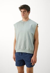 Grå uten ermer hettegenser med rund hals og ribbestrikket kant, kombinert med blå shorts. Materialet ser mykt og uformelt ut. Nike-logoen er synlig.