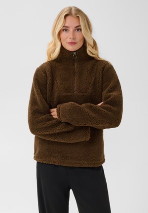 Femme aux longs cheveux blonds portant une veste polaire marron à fermeture éclair et un pantalon noir, debout les bras croisés devant un fond uni.