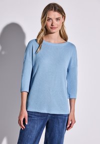 Helles blaues, strukturiertes Strickoberteil mit weitem Bootsausschnitt und 3/4-Ärmeln, kombiniert mit dunkelblauen Jeans. Einfaches und lässiges Design.